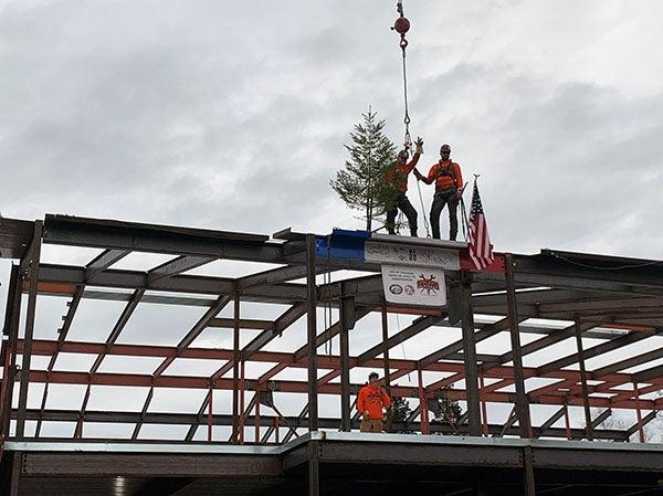 final beam in place with two people standing on it; it has a US flag, an evergreen tree, and a banner on it