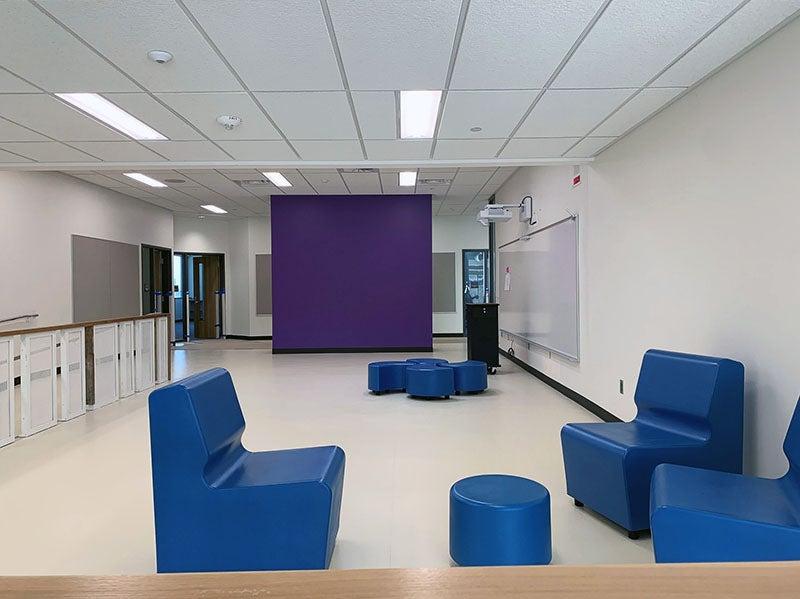 a group area has seating clusters, a learning wall, and a teacher station with a purple wall separating it from the hallway