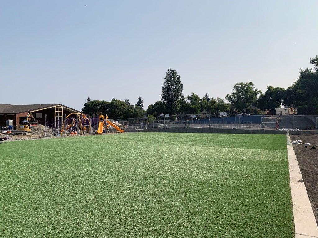 a broad expanse of artificial turf with a baskeball play are behind it